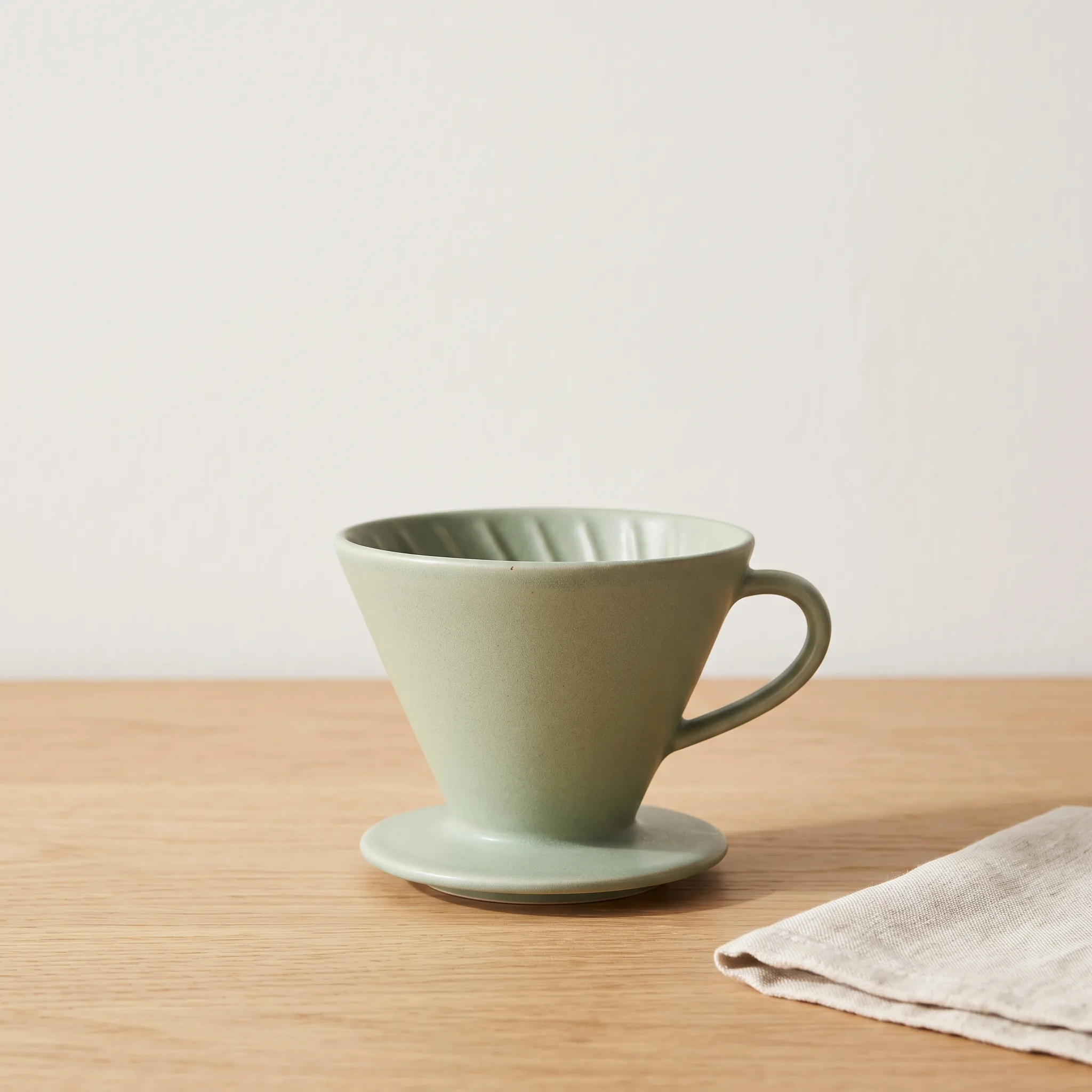 Nano Banana 2.1 e-commerce shot of a sage-green matte ceramic pour-over dripper on light oak, with open space above