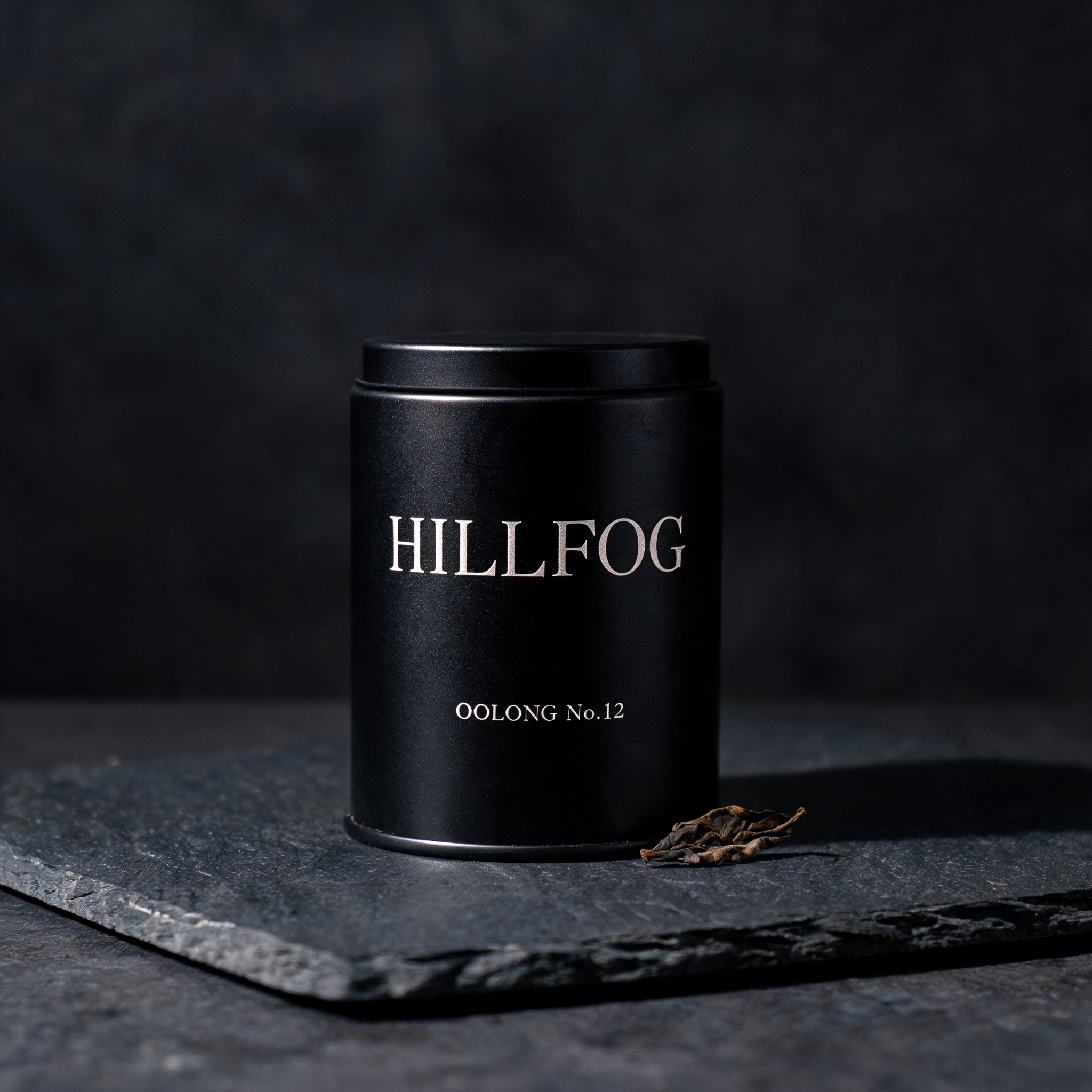 Studio photo of a matte-black tea tin labeled HILLFOG OOLONG No.12 on a slate surface with a single dried tea leaf beside it