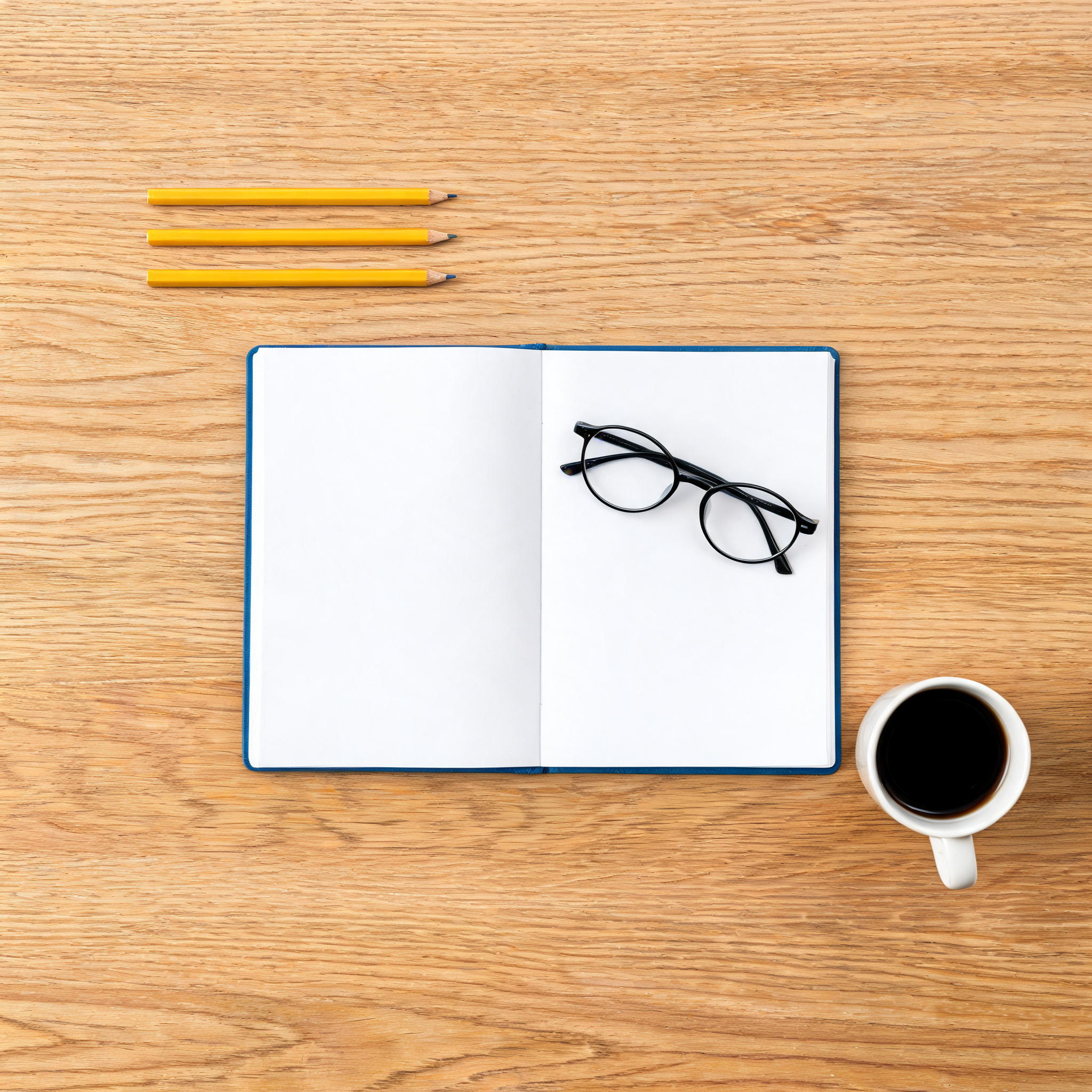Overhead flat-lay of a desk with exactly three yellow pencils top-left, an open blue notebook in the center, round glasses on its right page, and a mug of black coffee bottom-right